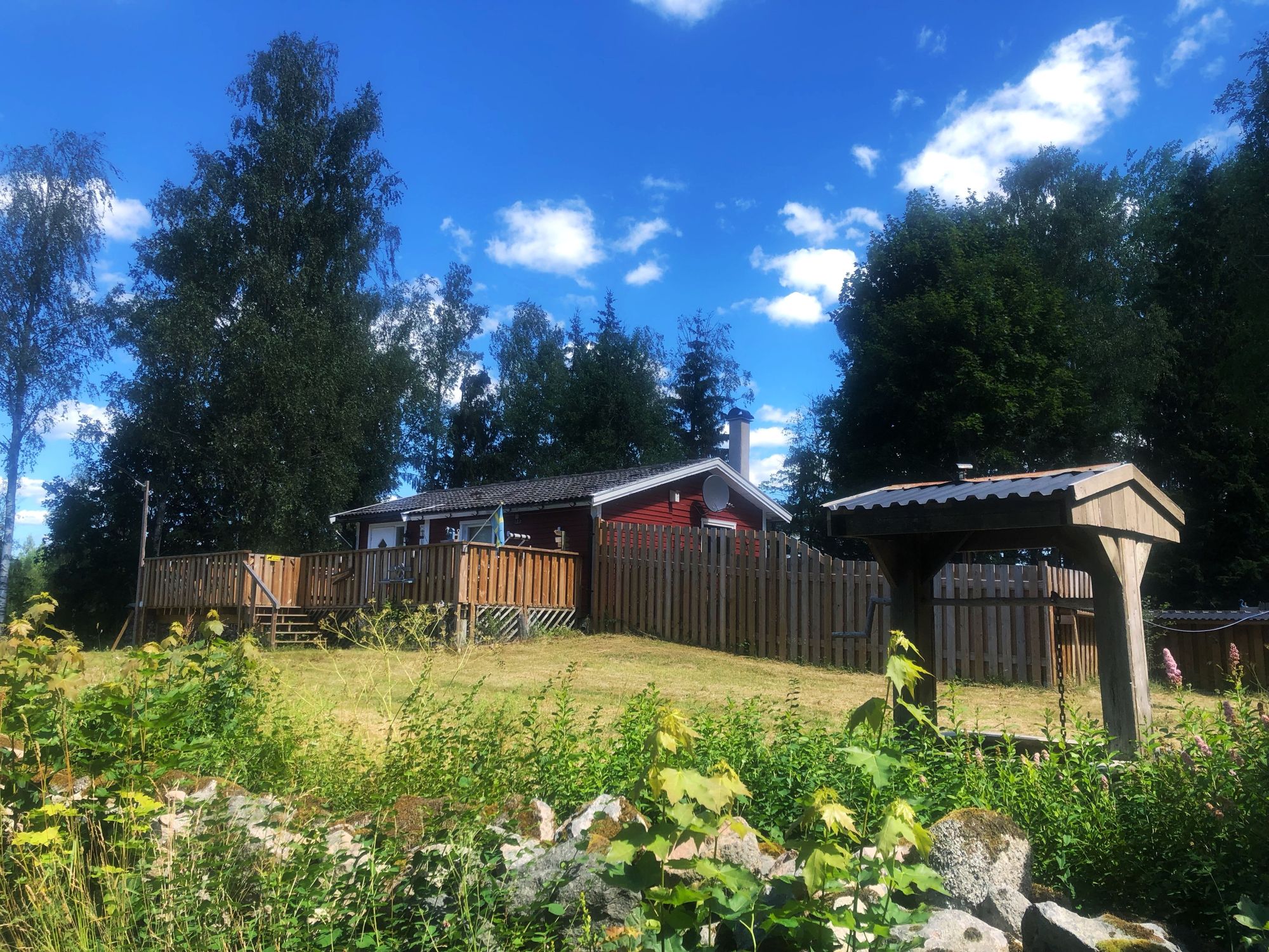 Das rote Ferienhaus mit Holzterrasse, Zaun und Brunnen, umgeben von Birken und Fichten unter blauem Sommerhimmel